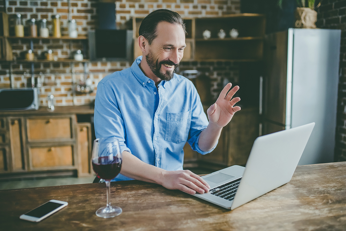 Un homme, devant son ordinateur portable, salue de la main un interlocuteur en visio-conférence. Il a un verre de vin posé à côté de lui, sur la table.